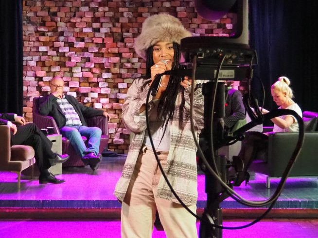 Rap-artisti Yeboyah laulaa mikrofoni kädessä kameran edessä, taustalla G Livelabin lavalla, jossa istumassa keskustelijoita.